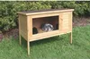 A wooden rabbit hutch with two rabbits inside, placed on a grassy lawn with bushes in the background. Voir les images du produit