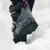 A close-up shot of a person's foot wearing a black and gray winter boot with a pink accent, stepping into snow. Podgląd zdjęcia produktu