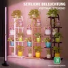 A vertical LED grow light stands next to multiple tiered plant stands filled with various potted plants. Náhľad obrázku produktu