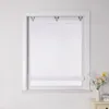 A white Roman blind is installed in a white window frame against a white wall. A small vase with flowers sits on a bedside table to the right of the window. Produktbild-Vorschau