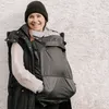 A woman wearing a black beanie and a black vest over a cream sweater, with a black baby carrier cover attached to the vest. Náhled obrázku produktu