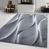 A modern abstract design rug with a grey and white wavy pattern is shown on a light wooden floor, with a fireplace and logs in the background. Produktbild-Vorschau