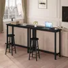 A long, narrow black bar table is placed behind a sofa, with two black bar stools in front of it. A laptop, a cup, and a small vase of flowers are on the table. Náhľad obrázku produktu