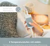 A person's hand holding a heating pad controller, with a cozy blanket and mug in the background. Produktbild-Vorschau