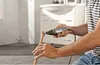 A person is using a Dremel tool to cut a copper pipe in a bathroom setting. Produktbild-Vorschau