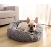 A French Bulldog is resting in a round, fluffy gray dog bed placed on a wooden floor next to a sofa. Anteprima dell’immagine prodotto