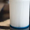 A close-up of a white pleated filter cartridge with a blue base, resting on a reflective black surface. Produktbild-Vorschau
