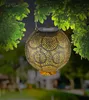 A decorative solar lantern hangs from a tree branch, illuminated by its internal light. Produktbild-Vorschau