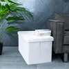 A white fabric storage ottoman sits next to a dark gray sofa, with a potted plant to the left and a tiled wall in the background. Podgląd zdjęcia produktu