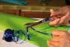 Hands are shown cutting blue ribbon with scissors on a green surface, with a roll of ribbon nearby. Náhled obrázku produktu