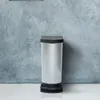 A silver and black trash can stands against a textured gray wall. Voir les images du produit