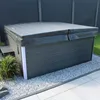 An outdoor hot tub covered with a dark gray thermal cover, situated on a gravel and tiled patio area, with a dark gray fence in the background. Podgląd zdjęcia produktu