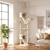 A tall cat tree is set up in a living room with a sofa and a plant. Produktbild-Vorschau