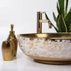 A close-up shot of a decorative washbasin with gold accents and floral patterns, accompanied by a gold soap dispenser and a toothbrush holder. A snake plant is visible in the background. Produktbild-Vorschau