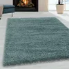 A shaggy blue rug is laid out on a light-colored wooden floor in front of a fireplace. A blue ottoman with a beige throw is visible to the left, and a large window with a snowy landscape is in the background. Produktbild-Vorschau