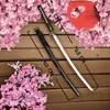 A katana sword and its scabbard are displayed on a wooden surface, surrounded by pink cherry blossoms. Measurements are indicated with lines and text. Produktbild-Vorschau