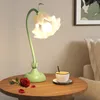 A flower-shaped table lamp is placed on a wooden table next to a cup of coffee and a red rose. Produktbild-Vorschau