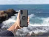 A hand holding a phone case with a mount, with the ocean and rocks in the background. Produktbild-Vorschau
