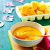 A bowl of mango puree with a spoon, next to a tray of mango chunks, with decorative letters in the background. Podgląd zdjęcia produktu