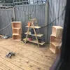 An outdoor patio with wooden decking, a grey wooden fence, and various wooden crates arranged as shelving and storage. There are also dumbbells and plants visible. Náhled obrázku produktu