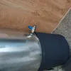 A close-up view of a stainless steel water heater with a black insulating sleeve and a blue valve, placed next to a wooden surface and a concrete floor. Produktbild-Vorschau