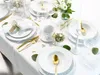 A beautifully set table with white porcelain dinnerware and gold cutlery, decorated with greenery and a white candle. Produktbild-Vorschau