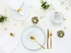 A beautifully set table with white plates, gold cutlery, and small floral arrangements. Produktbild-Vorschau