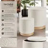 Two cream-colored, bouclé-covered storage ottomans with wooden bases are displayed in a modern living room setting. A plant in a dark pot is visible in the background. Náhled obrázku produktu