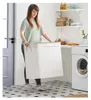 A woman is holding a large white laundry basket in a laundry room with a washing machine and shelves. Podgląd zdjęcia produktu