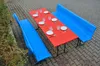 A red table with blue benches set up outdoors on a paved area, with grass on the side. Produktbild-Vorschau