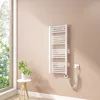 A white electric towel radiator mounted on a light pink wall next to a large window. A small white side table with a green plant in a white vase is to the left of the radiator. Produktbild-Vorschau