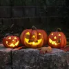 Three illuminated jack-o'-lanterns sit on a stone wall at dusk, with fallen leaves scattered around them. Anteprima dell’immagine prodotto