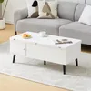 A white coffee table with a closed cabinet door and a magazine on top, placed in front of a gray sofa with decorative pillows. A cup and pastries are on the table. Produktbild-Vorschau