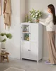 A woman is placing a vase of flowers on top of a white bathroom cabinet. The cabinet has four doors and is filled with towels and jars. Produktbild-Vorschau