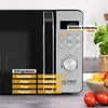 A close-up view of a microwave oven's control panel and a portion of its door, with food items and a plant in the background. Produktbild-Vorschau