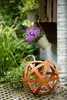 A decorative metal sphere sits on a patterned surface in a garden setting, with plants and hanging baskets in the background. Produktbild-Vorschau