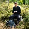 A young man is sitting on a tree stump in a forest, looking at a map. A backpack and hiking poles are next to him. Anteprima dell’immagine prodotto