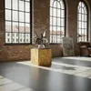 A modern loft-like interior with large windows, brick walls, and a concrete floor, featuring a sculpture on a wooden pedestal and a leather armchair. Produktbild-Vorschau