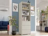A tall, narrow bookshelf with three drawers is shown in a well-lit room with blue walls. A plant is in the foreground, and other furniture and decor are visible in the background. Náhled obrázku produktu