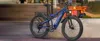 A blue electric mountain bike is parked on a sidewalk in front of a cafe. Produktbild-Vorschau