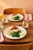 Two plates of food are set on a wooden table, with glasses of rosé wine and a vase of flowers. Anteprima dell’immagine prodotto