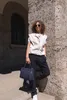 A woman stands on steps holding a navy blue handbag, leaning against a stone wall. Voir les images du produit