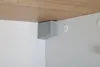 A close-up view of a corner of a cabinet or shelf unit, showing a gray corner protector under a wooden shelf and against a white wall. Podgląd zdjęcia produktu
