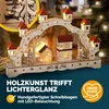 A wooden Christmas decoration with LED lights, featuring houses and trees, set against a blurred background of a fireplace and Christmas tree lights. Náhled obrázku produktu