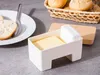 A butter dish with butter and a lid sits on a gray surface next to a plate with bread and a basket of rolls. Podgląd zdjęcia produktu