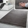 A soft, gray rug is shown in a modern living room setting, with a white sofa and a close-up inset of a hand pressing into the rug to demonstrate its plushness. Produktbild-Vorschau