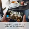 A person holding a smartphone displaying a security camera feed of a dog inside a home, with coffee and a pastry on a table in the background. Anteprima dell’immagine prodotto