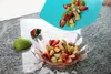 A fruit salad is being prepared, with chopped fruits being poured from a blue cutting mat into a glass bowl. A whole apple and strawberry are on the counter. Produktbild-Vorschau