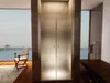 A modern bathroom with a frosted glass shower door, a chair by the window overlooking the ocean, and a wooden bench. Produktbild-Vorschau