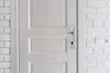 A close-up view of a white door with a silver handle and lock, next to a white brick wall with a power outlet. Náhled obrázku produktu